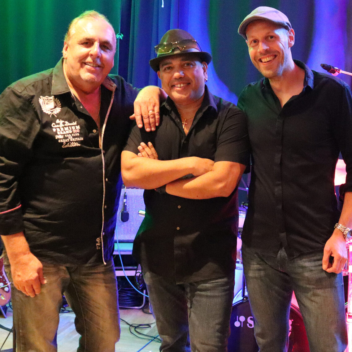 Drei Mäner im schwarzen Hemd stehen auf der Bühne und Lächeln in die Kamera. Im Hintergrund sieht man ein Schlagzeug und eine E-Gitarre.