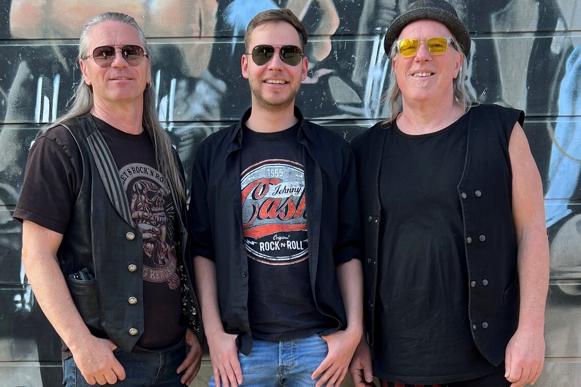 Links Mann mit langen grauen Haaren und Pilotensonnenbrille, Mitte Mann mit kurzen braunen Haaren und Pilotensonnenbrille, rechts Mann mit schulterlangen grauen Haaren, schwarzem Hut und Sonnenbrille mit gelben Gläsern.