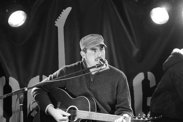 BEN’TAGON sitzt an der Gitarre und singt in ein Mikro. Das Bild hat einen schwarz weiß Filter und findet auf einer Bühne statt.