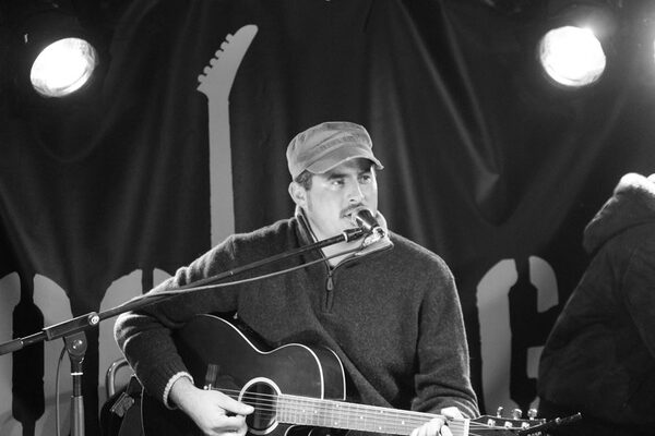 BEN’TAGON sitzt an der Gitarre und singt in ein Mikro. Das Bild hat einen schwarz weiß Filter und findet auf einer Bühne statt.