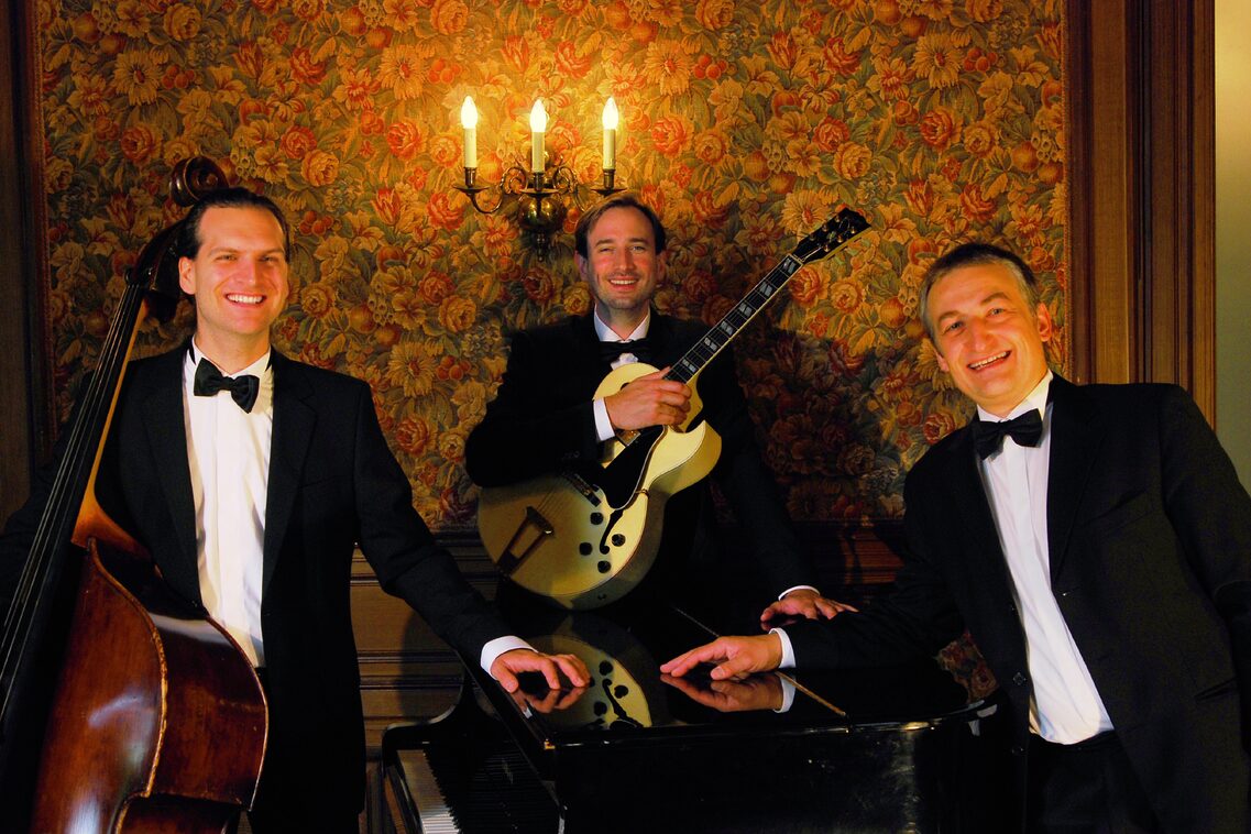 Група в стая със стари тапети с флорални мотиви и стенна светлина, всички в черни костюми с бели ризи и черни папийонки. Вляво мъж с бас китара, в средата мъж с електрическа китара, а вдясно мъж на роял. Стаята е много старомодна.