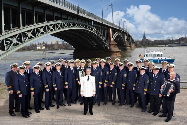Der Chor steht in typischer Seemannsbekleidung vor einer Brücke. Alle lachen in die Kamera. In der Mitte steht eine Frau, die anders gekleidet ist. Rechts hält ein Mann ein Akkordion.