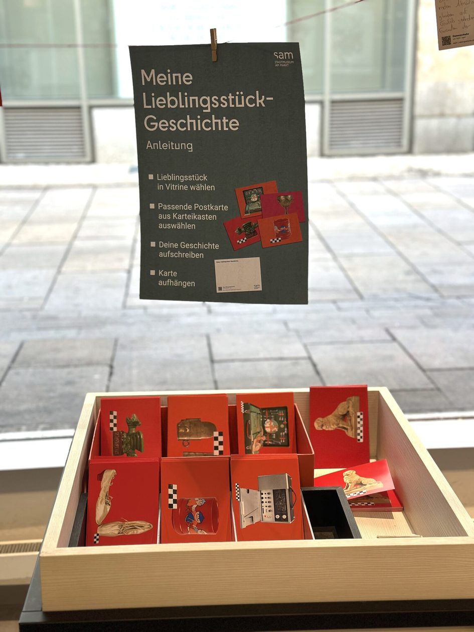 Ein Foto von einem Plakat, das mit der Aufforderung „Postkarten zu beschriften“ an einer Wäscheleine hängt, und verschiedene Postkarten.