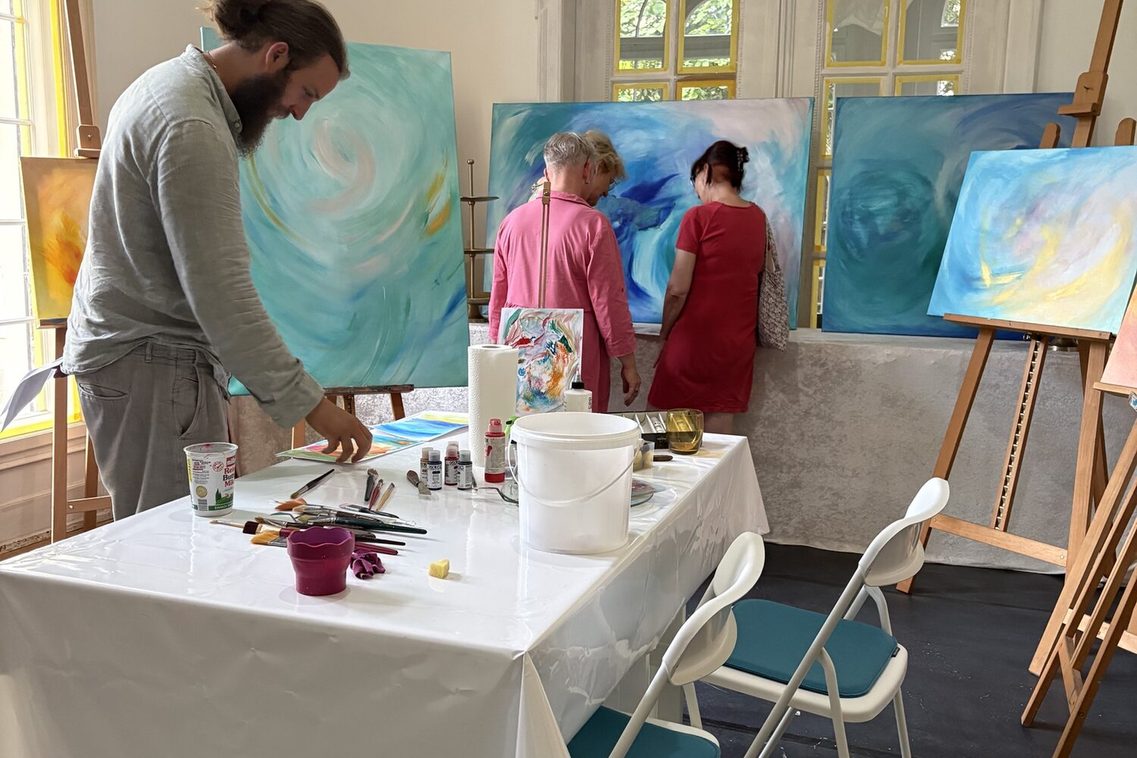 im Atelier, Tische mir Malutensilien vorne, Staffeleien und drei Personen im Hintergrund