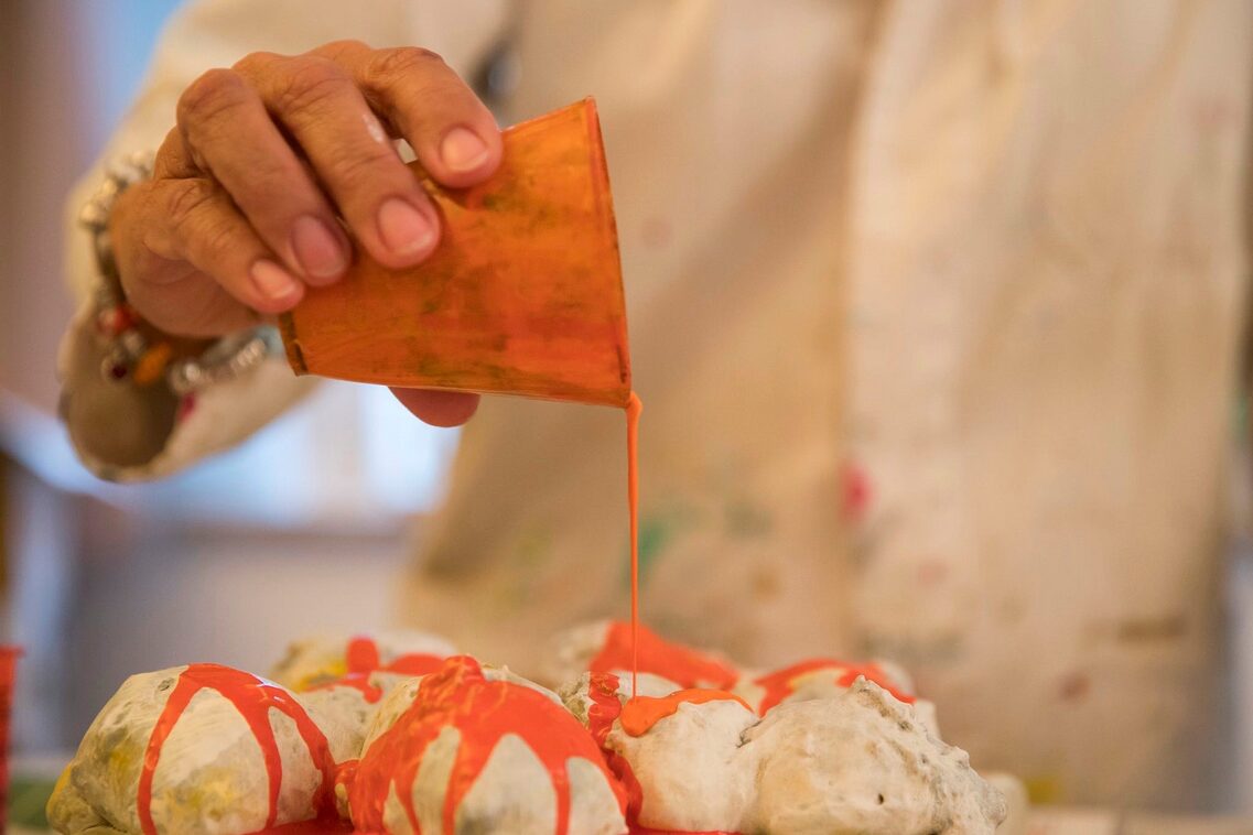 eine Hand gießt orangene Farbe aus einem Becher auf naturweiße eikugelförmige Objekte