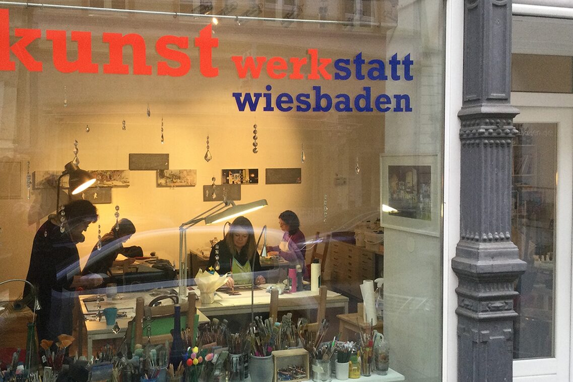 Blick durch die Fensterfront in das Atelier. Maluntensilien wie Pinsel und Stifte, weiter im Raum bei Licht vier Personen, die künstlerisch arbeiten.