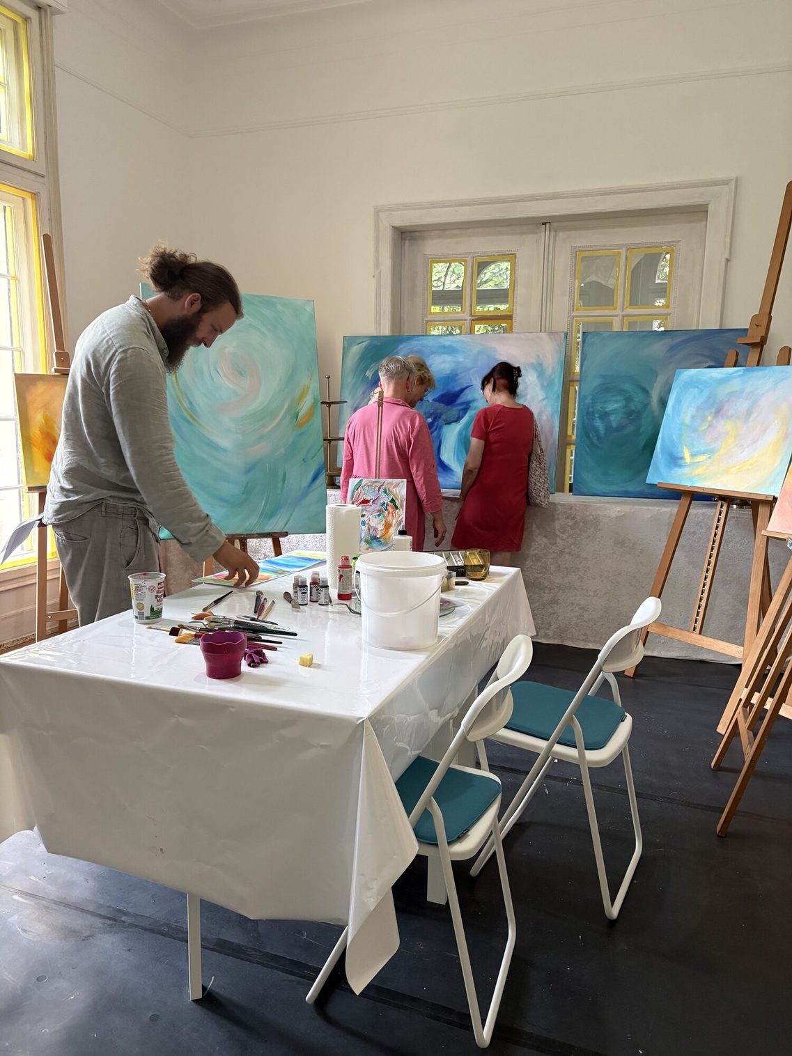 In the studio: tables with painting supplies in the foreground, easels, and three people in the background.