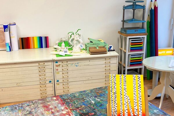 Blick in ein Atelier mit Schubladenschrank und bunter Tischunterlage und Kunstwerk im Vordergrund