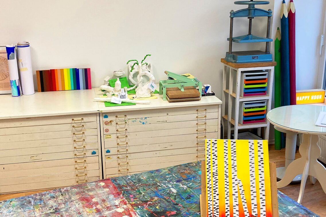 Blick in ein Atelier mit Schubladenschrank und bunter Tischunterlage und Kunstwerk im Vordergrund