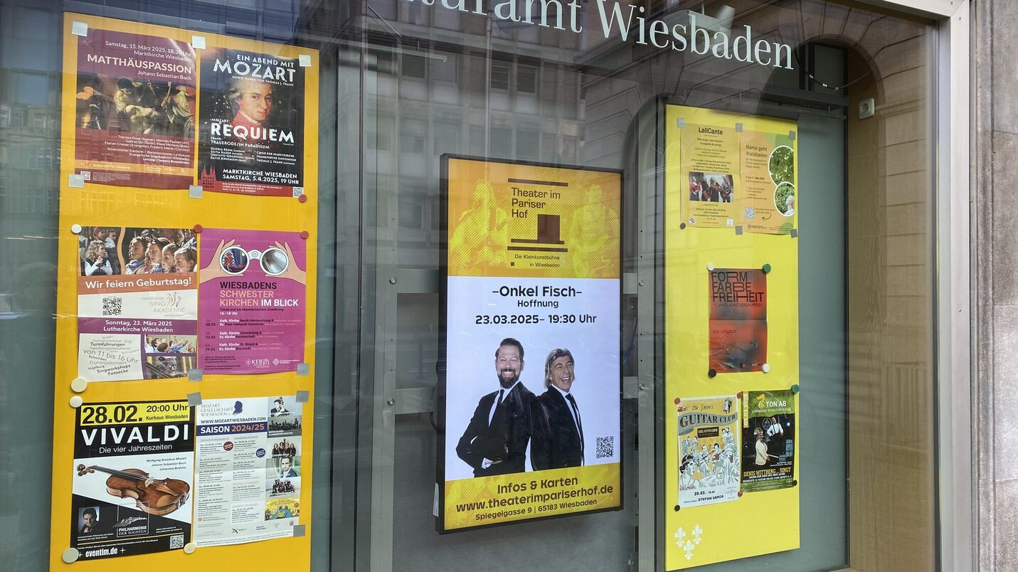 Okno wystawowe biura kultury z plakatami wydarzeń kulturalnych, wyśrodkowane na cyfrowej steli z informacjami o wydarzeniach w teatrze w Pariser Hof.