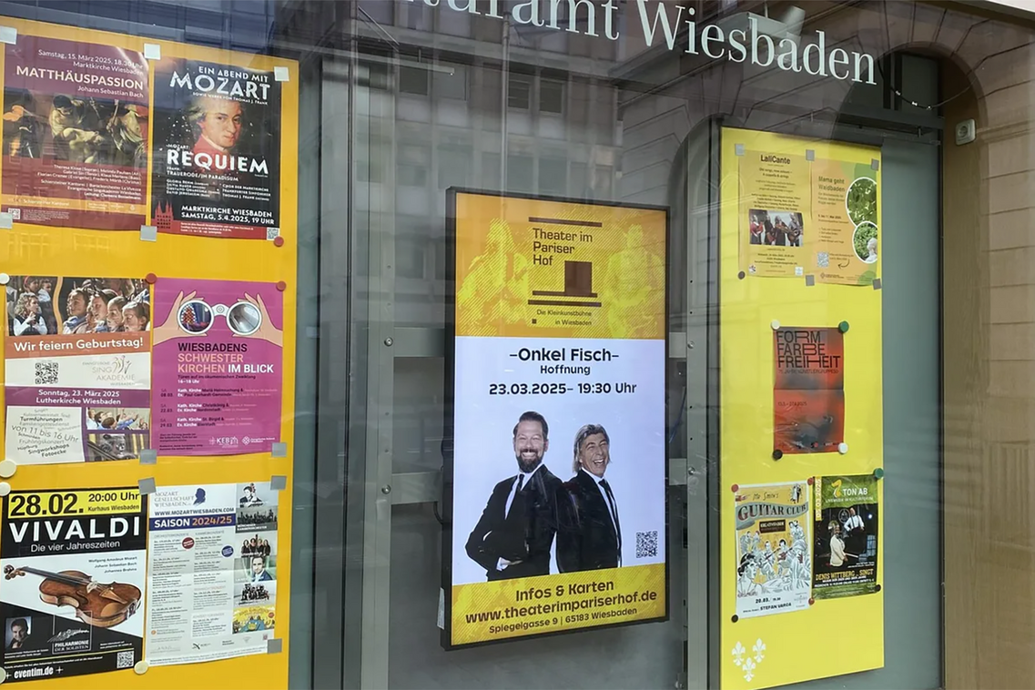 Schaufenster des Kulturamtes mit Postern von Kulturveranstaltungen, zentriert eine digitale Stele mit Veranstaltungshinweisen des Theaters im Pariser Hof
