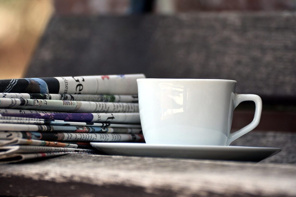 A white coffee cup with saucer stands next to a folded stack of newspapers