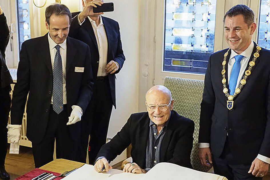 Трое мужчин в темных костюмах, мужчина в центре сидит и пишет.