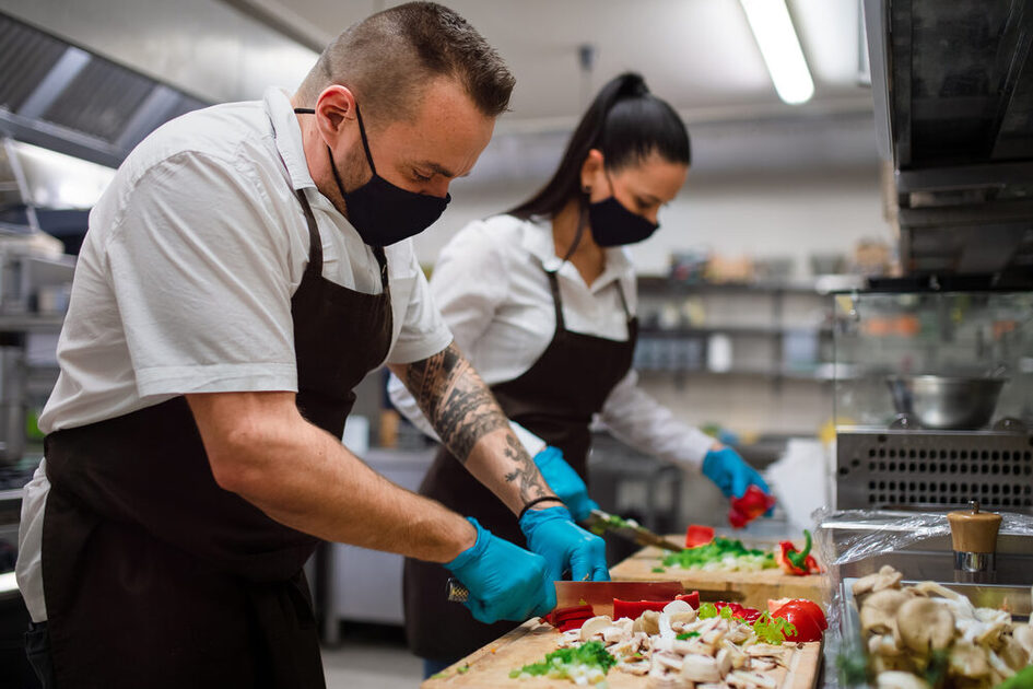 Das Bild zeigt einen Mann und eine Frau, die Gemüse schneiden in einer Großküche.