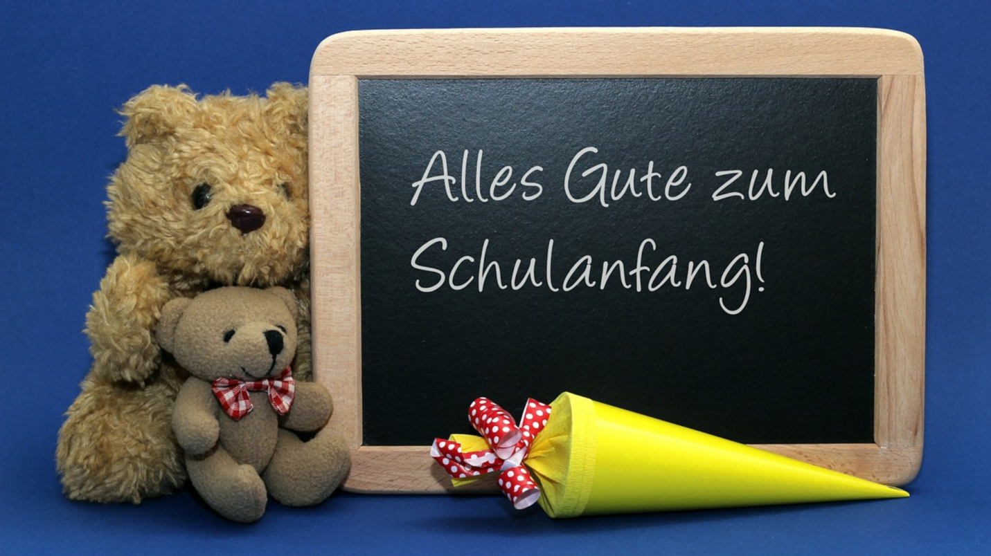 Tafel mit Text: alles Gute zum Schulanfang. Zu sehen zwei Stoffbären links der Tafel und im Vordergrund eine gelbe Schultüte