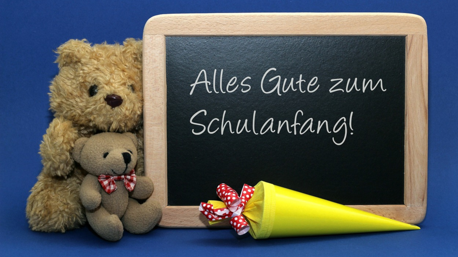 Tafel mit Text: alles Gute zum Schulanfang. Zu sehen zwei Stoffbären links der Tafel und im Vordergrund eine gelbe Schultüte