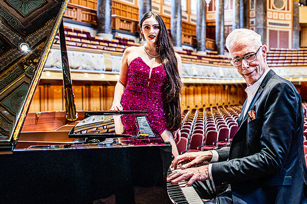 La donna è in piedi davanti al pianoforte a coda, l'uomo si siede davanti ad esso e suona.