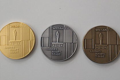 Es sind drei Medaillen auf grauem Untergrund zu sehen in Gold, Silber Bronze. Darauf steht Wiesbaden Landeshauptstadt.