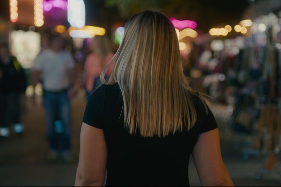 Frau mit langen blonden Haaren geht vor dem Betrachter her durch eine Menschenmenge in Dunkelheit.