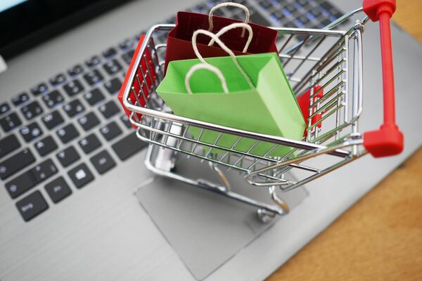 Miniatur-Einkaufswagen steht auf der Tastatur eines Laptops. In dem Einkaufswagen befinden sich kleine Tüten.