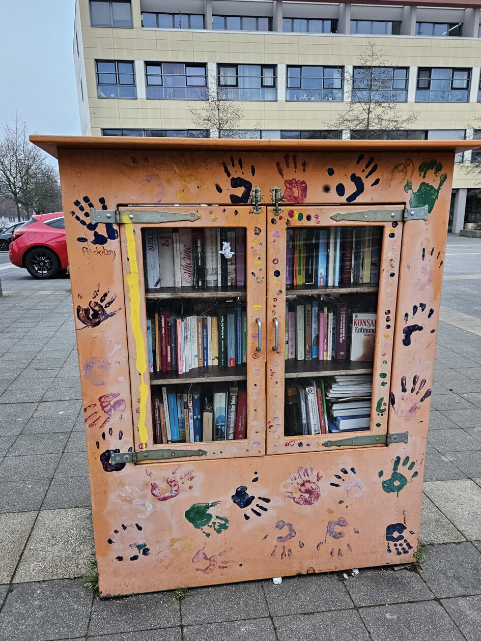 A cupboard stands outside in a square. It is painted apricot and decorated with colorful handprints. There are books in the cupboard.