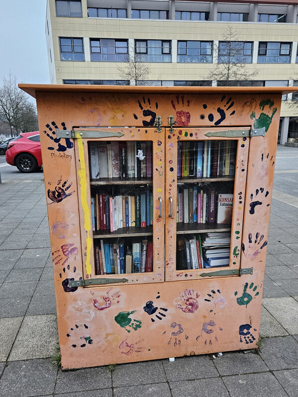 Ein Schrank steht draußen auf einem Platz. Er ist apricotfarben gestrichen und mit bunten Handabdrücken verziert. In dem Schrank sind Bücher zu sehen.