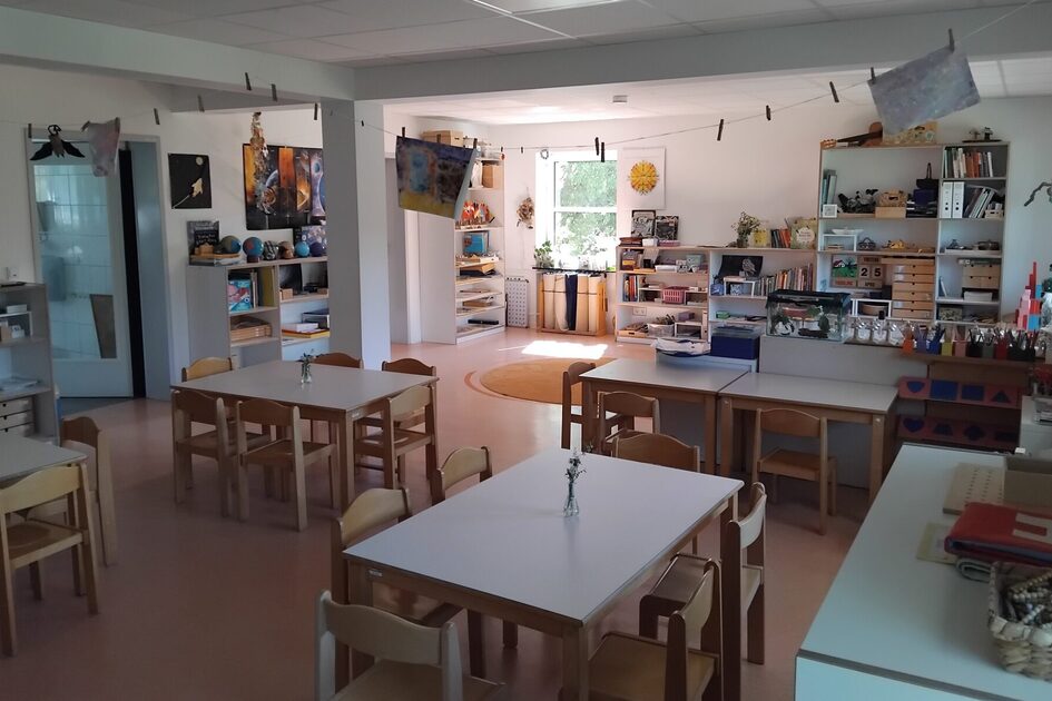 Hier sehen Sie den Gruppenraum mit Tischen und Stühlen. An der hinteren Wand stehen Regale