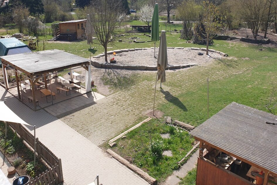 Das Außengelände der Kindertagesstätte mit Hütten, einem großen runden Sandspielbereich und großen Sonnenschirmen