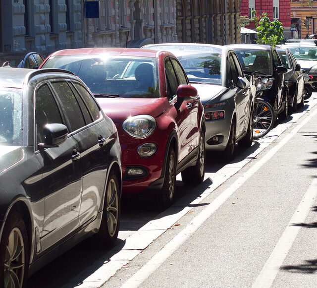 Parkende Autos an einem Gehweg, im Hintergrund historische Häuser.