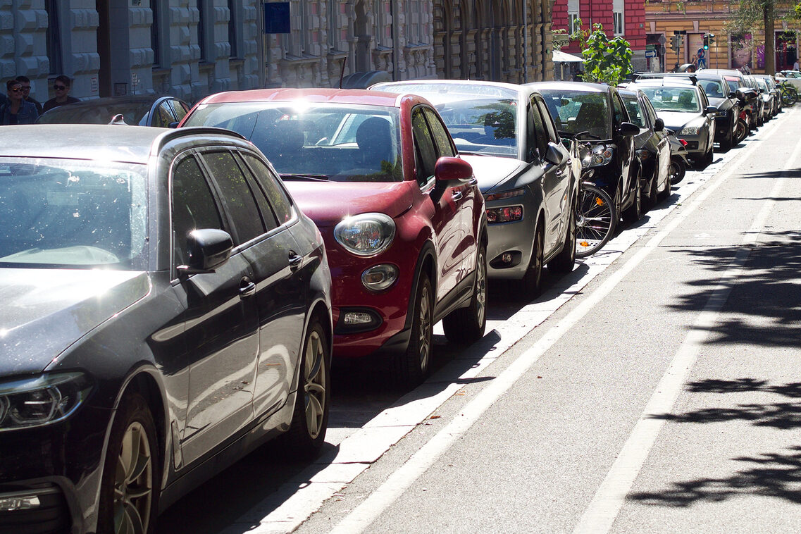 Parkende Autos an einem Gehweg, im Hintergrund historische Häuser.