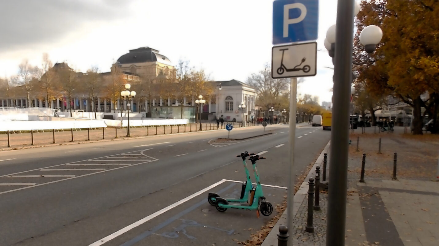 Das Bild zeigt eine Straße mit einer blau markierten Parkfläche für E-Scooter, gekennzeichnet durch ein Schild mit einem „P“ und einem Roller-Symbol. Zwei türkisfarbene E-Scooter stehen dort. Im Hintergrund sind das Theater der Stadt Wiesbaden, die Weihnachtseisbahn und herbstliche Bäume zu sehen. Der Himmel ist bewölkt, und auf dem Boden liegen einige Blätter.