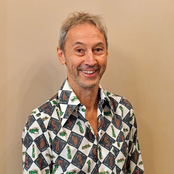 Person with gray hair wears a shirt with a pattern of black and white diamonds, numbers and green figures.