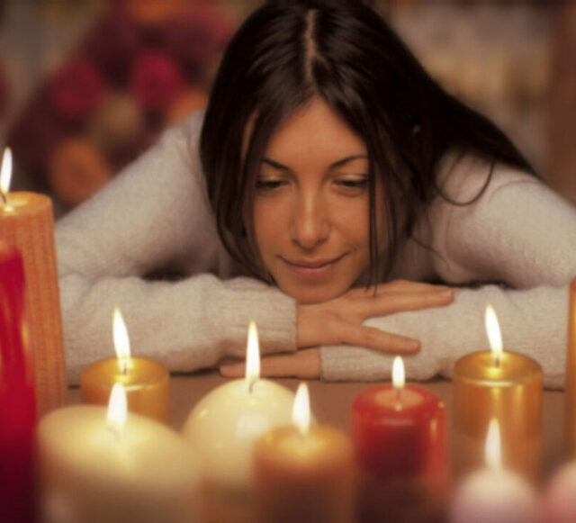En arrière-plan, on voit une femme aux longs cheveux bruns portant un pull en laine blanc, ses mains sont posées l'une sur l'autre sur une surface et son menton est posé sur ses mains, son regard est dirigé vers le bas. Au premier plan se trouvent plusieurs bougies allumées en cire blanche, dorée et rouge.