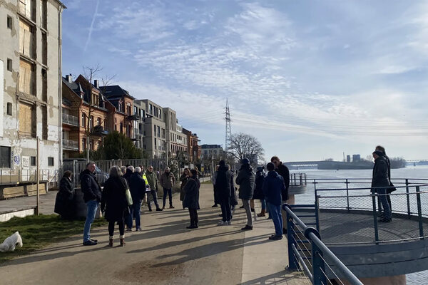 Das Foto zeigt eine Menschengruppe von etwa 25 Personen, die an einem Frühlingstag auf der Promenade des Rheinufers vor einem kaputt aussehenden Gebäude stehen. Die Menschen sind mit einander im Gespräch. Die Umgebung des beschädigten Gebäudes ist malerisch: Man kann weit den Fluss hinauf schauen. Der Himmel leuchtet in einem klaren, strahlenden Blau. Die Häuser im Bildhintergrund sind intakt. Ein einprägsames Bild.