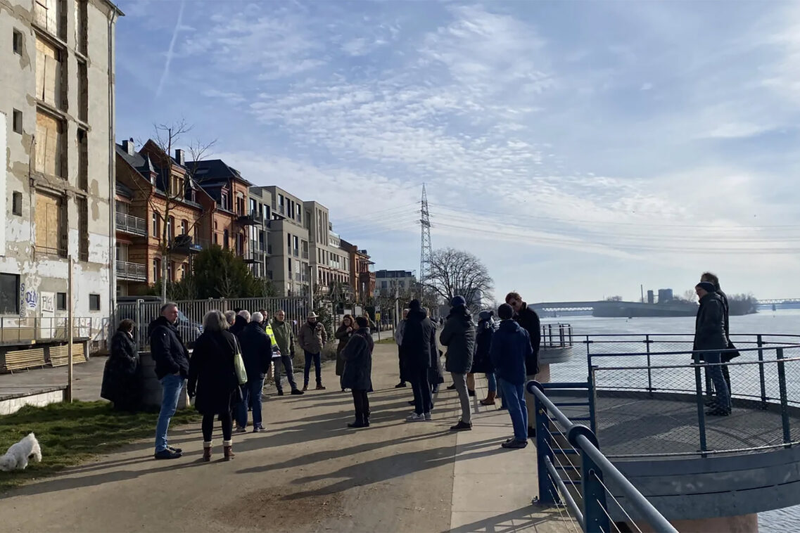Das Foto zeigt eine Menschengruppe von etwa 25 Personen, die an einem Frühlingstag auf der Promenade des Rheinufers vor einem kaputt aussehenden Gebäude stehen. Die Menschen sind mit einander im Gespräch. Die Umgebung des beschädigten Gebäudes ist malerisch: Man kann weit den Fluss hinauf schauen. Der Himmel leuchtet in einem klaren, strahlenden Blau. Die Häuser im Bildhintergrund sind intakt. Ein einprägsames Bild.