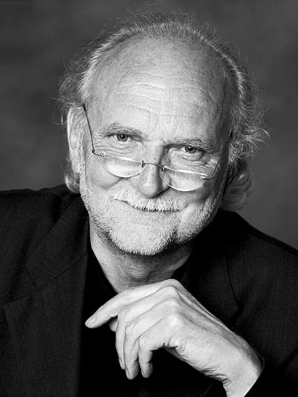 Professor Roland Burgard can be seen in the black and white picture. He wears frameless glasses and smiles at the camera in a friendly manner with a slightly mischievous expression. Mr. Burgard leans forward slightly, his hand is clearly visible. His posture and facial expression are inviting and friendly. He is wearing dark clothing, which goes well with the black and white aesthetic of the picture.
