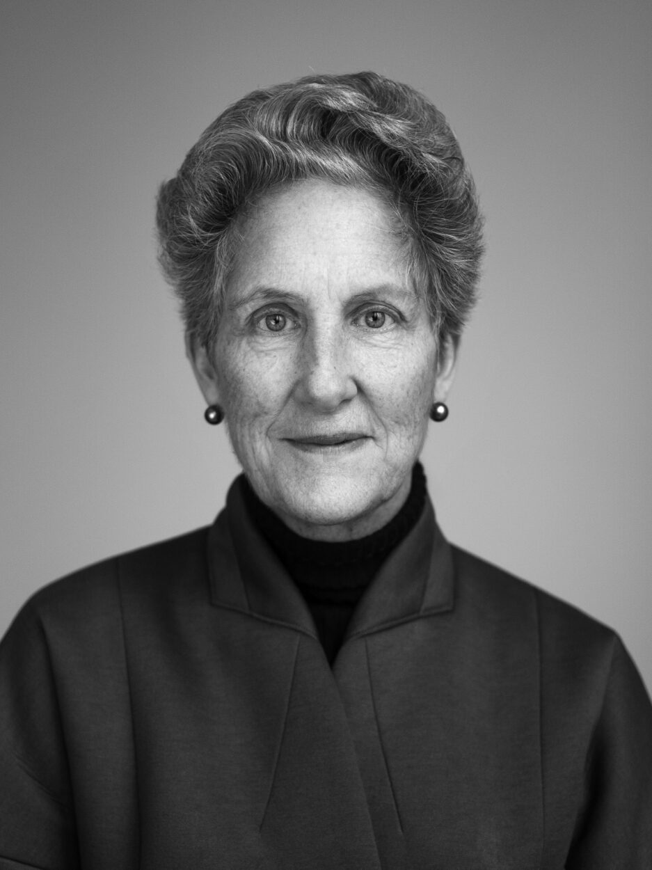 Das Bild zeigt eine ältere Frau mit kurzen, gewellten Haaren, die ordentlich nach hinten frisiert sind. Sie hat feine Gesichtszüge mit markanten Wangenknochen und trägt kleine runde Ohrringe. Ihr Ausdruck ist freundlich und würdevoll, mit einem leichten Lächeln. Sie trägt eine dunkle, elegante Jacke mit einem hohen Kragen über einem schwarzen Rollkragenpullover. Der Hintergrund ist schlicht und neutral, was den Fokus auf ihr Gesicht lenkt. Die Aufnahme ist in Schwarz-Weiß gehalten, was den Kontrast und die Details ihrer Gesichtszüge betont.
