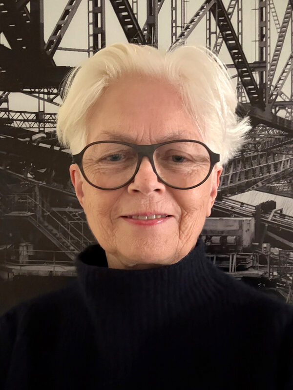 Fotoğrafta Profesör Sophie Wolfrum bir selfie çekerken ve neşeyle gülümserken görülüyor. Sanayi tarihinden bir sahneyi tasvir eden bir posterin önünde duruyor ve çarpıcı ama incelikli bir şekilde çözülen çelik kiriş yapılarını gösteriyor. Bayan Wolfrum beyaz saçlı, siyah, kavisli gözlükler takıyor ve siyah boğazlı bir kazak giyiyor. Neşeli gülümsemesi ve yaratıcı, şık kıyafetleri posterin endüstriyel temasıyla ilginç bir tezat oluşturuyor.