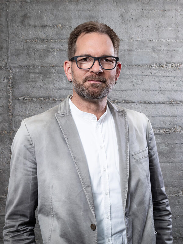 Portraitfoto in Farbe von Herrn Gleim. Herr Gleim steht vor einer grauen Betonwand und blickt mit neutralem Gesichtsausdruck direkt in die Kamera. Er trägt ein weißes Hemd und darüber ein hellgraues Sakko. Er hat kurze dunkelblonde oder hellbraune Haare und trägt eine schwarze Brille mir großen, eckigen, leicht abgerundeten Gläsern. Er hat einen kurzen Vollbart und trägt kleine, silberne Ohrringe.