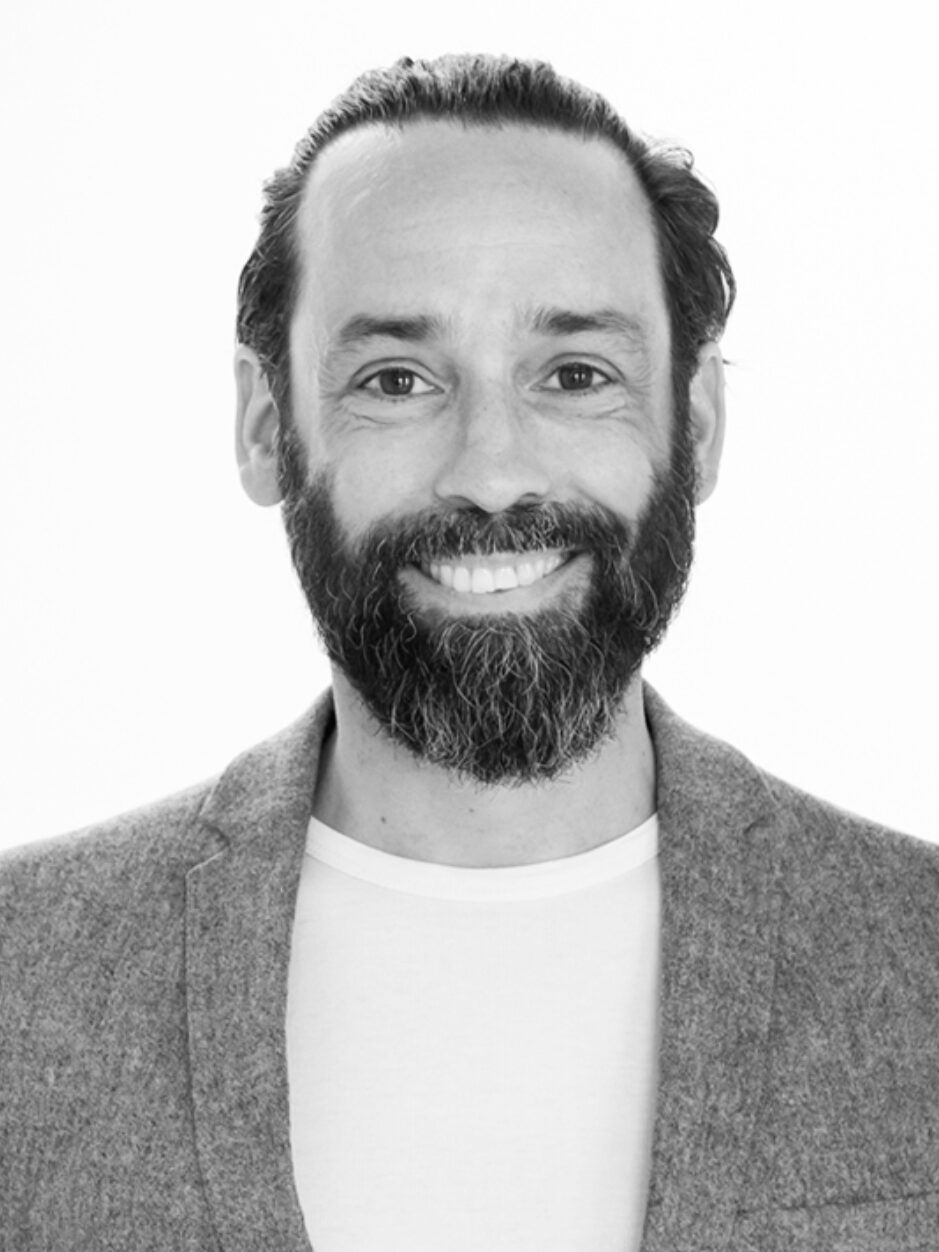 Portraitfoto in schwarz-weiß von Herrn Kist. Herr Kist befindet sich vor einem weißen Hintergrund. Er trägt ein Shirt und ein meliertes Sakko darüber. Herr Kist grinst freundlich in die Kamera. Er hat einen dunklen, gestutzten Vollbart mit vereinzelten grauen Haaren. Seine Haupthaar ist dunkel und glatt und nach hinten gebunden oder gekämmt. Er hat dunkle Augen.