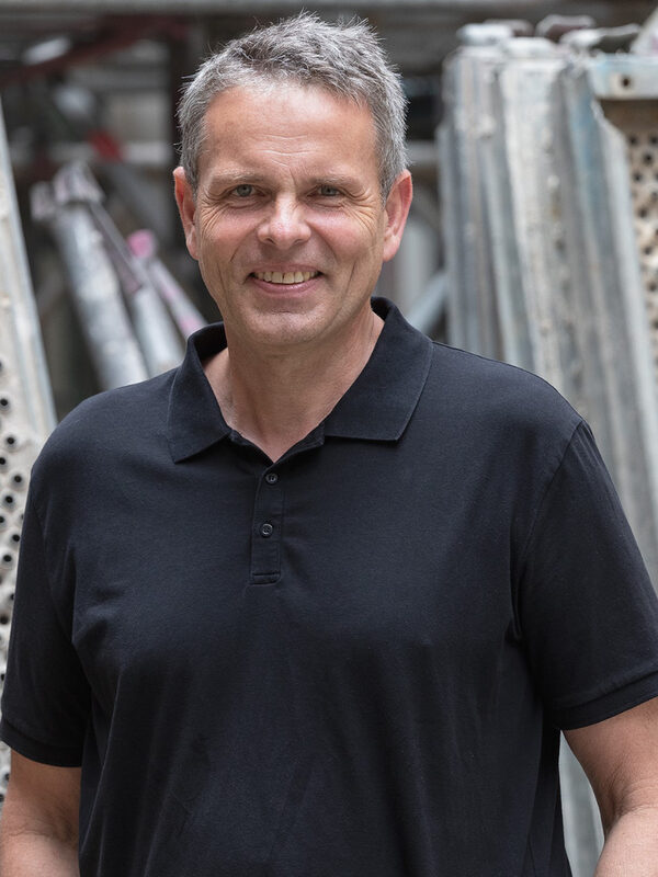 Ein Mann steht vor Einzelteilen eines Baugerüsts. Er trägt ein Polohemd und lächelt.