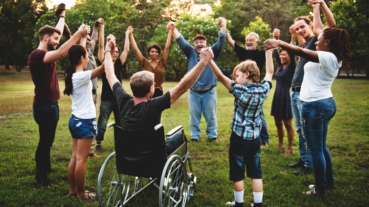 Menschen verschiedener Ethnien, verschiedener Geschlechter und verschiedenen Alters halten einander an den Händen und heben diese gemeinsam in die Höhe. Die Menschen bilden einen Kreis und befinden sich auf einer grünen Wiese mit Bäumen im Hintergrund. Eine der Personen sitzt im Rollstuhl. Die Menschen sind sommerlich gekleidet.