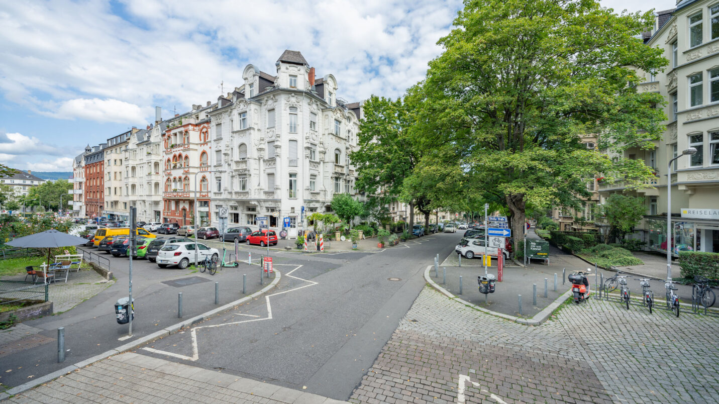 Man sieht eine große Kreuzung. Die Fläche besteht aus unterschiedlichen Pflastern und Asphalt. Rechts im Bild befinden sich Bäume, in der Straße links gibt es keine Bäume mehr, nur parkende Autos.