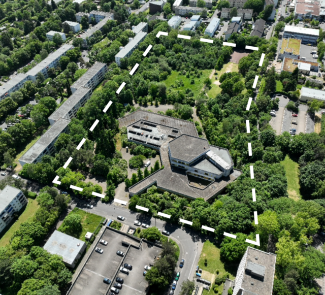 Ukośny widok z lotu ptaka na teren dawnej szkoły z białą przerywaną linią wokół granicy nieruchomości. Na zdjęciu dominują kolory szary i zielony. Można rozpoznać budynek szkoły w kształcie plastra miodu i wiele drzew wokół niego. Na skraju fragmentu obrazu znajdują się wielopiętrowe budynki mieszkalne i parking na pierwszym planie. Jest słonecznie.