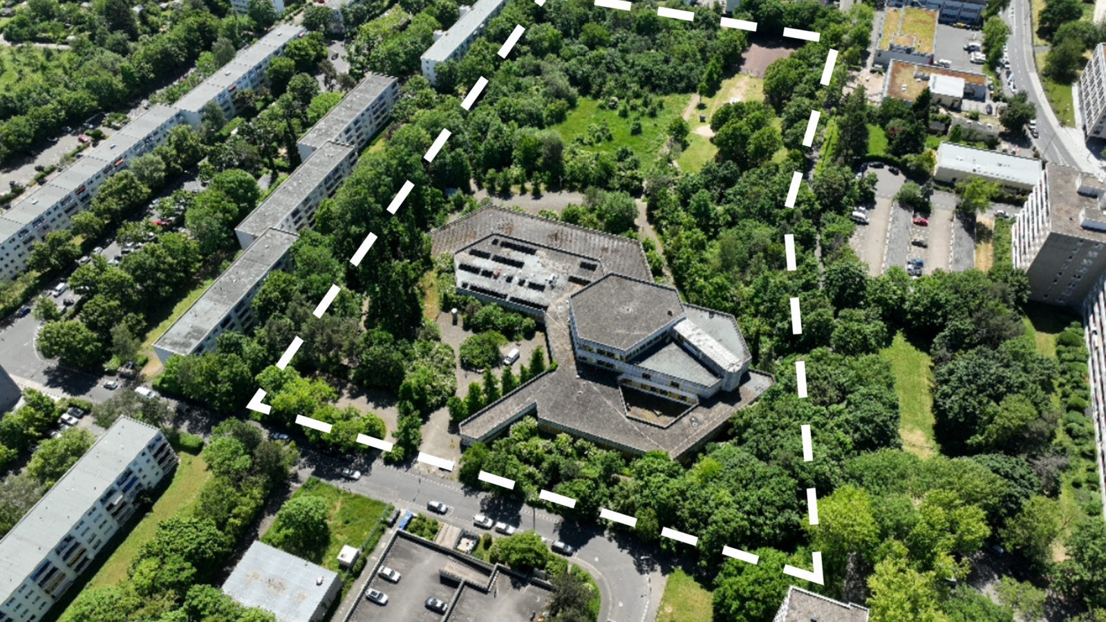 Ukośny widok z lotu ptaka na teren dawnej szkoły z białą przerywaną linią wokół granicy nieruchomości. Na zdjęciu dominują kolory szary i zielony. Można rozpoznać budynek szkoły w kształcie plastra miodu i wiele drzew wokół niego. Na skraju fragmentu obrazu znajdują się wielopiętrowe budynki mieszkalne i parking na pierwszym planie. Jest słonecznie.