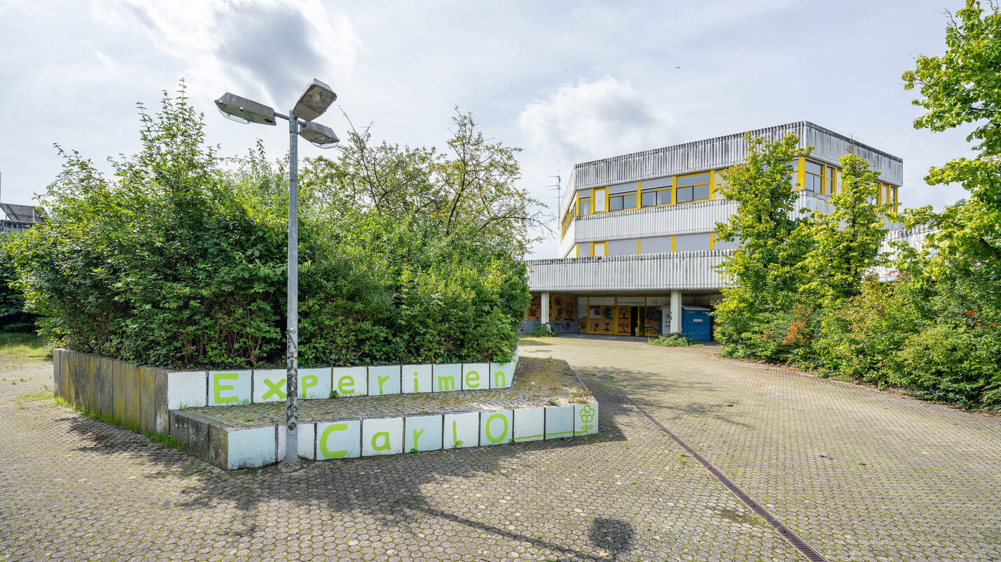 Das Bild zeigt ein Gebäude mit einer modernen, funktionalen Architektur. Die Fassade ist überwiegend grau mit gelben Fenstereinfassungen. Im Vordergrund befindet sich eine gepflasterte Fläche mit einer niedrigen, bepflanzten Mauer, auf der in grünen Buchstaben "Experimen Carlo" steht. Eine ältere Straßenlaterne mit zwei Leuchten wirft einen langen Schatten auf den Boden. Der Bereich wirkt etwas verlassen, mit Bewuchs an den Rändern. Im Hintergrund ist der Himmel mit einigen Wolken zu sehen.