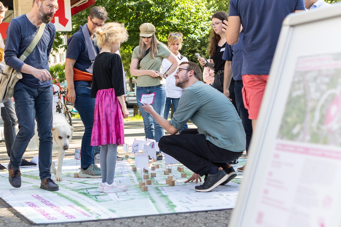 Verschiedene Menschen stehen auf einem Teppich. Ein Mann zeigt einem Kind einen Zettel.
