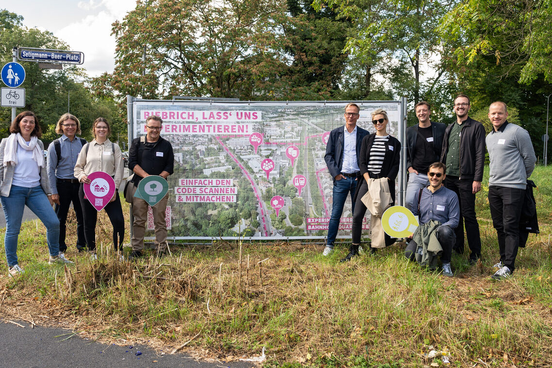 Eine Gruppe von Frauen und Männern steht lächelnd vor und neben einem Plakat auf einer Rasenfläche.