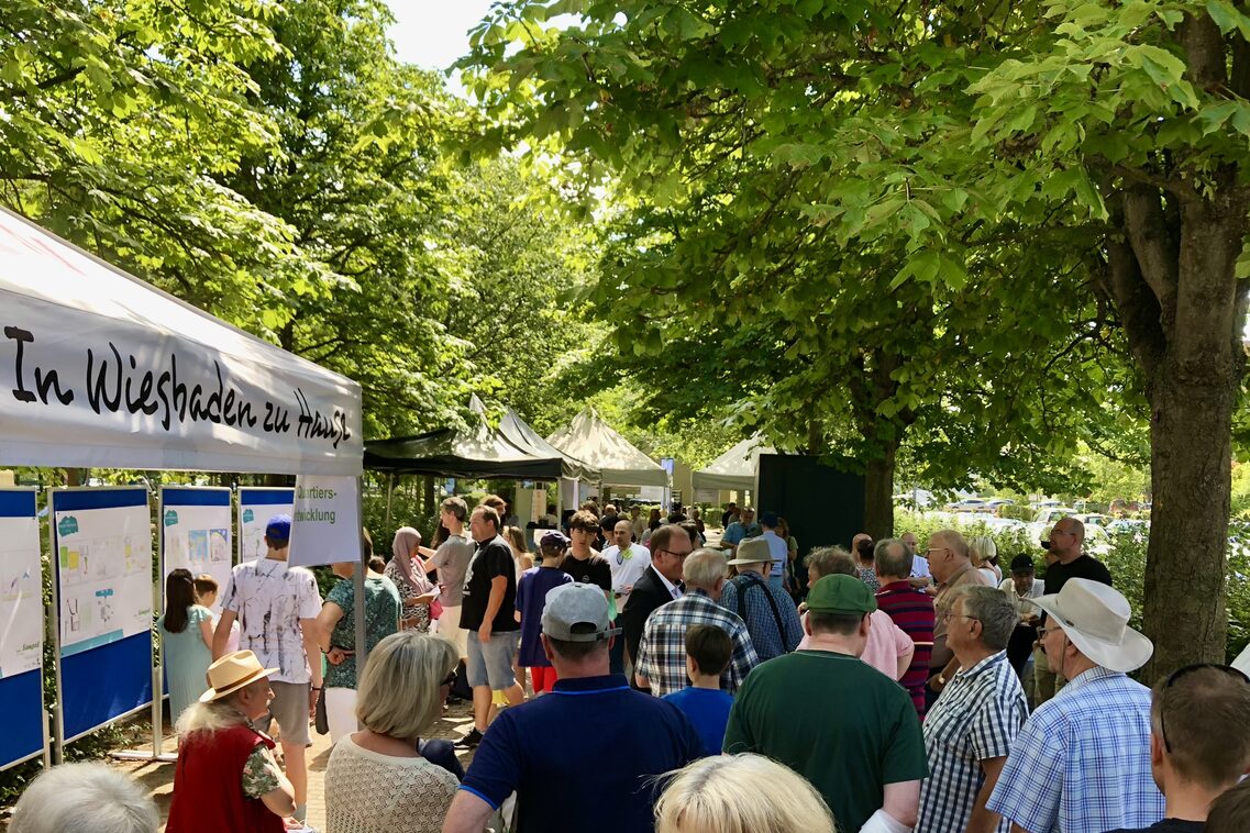 Viele Menschen aller Altersgruppen stehen zusammen, es sind verschiedene Pavillons und Informationstafeln zu sehen.