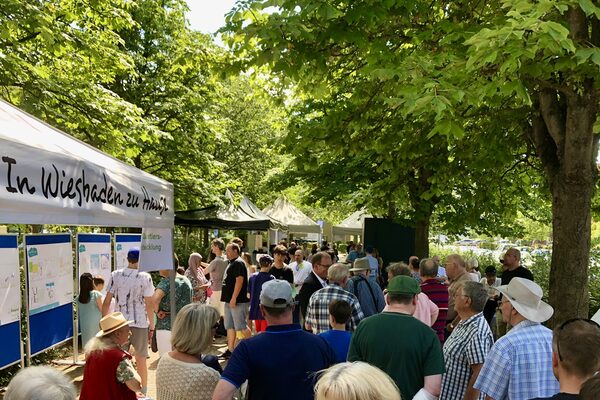 Many people of all ages stand together, there are various pavilions and information boards.